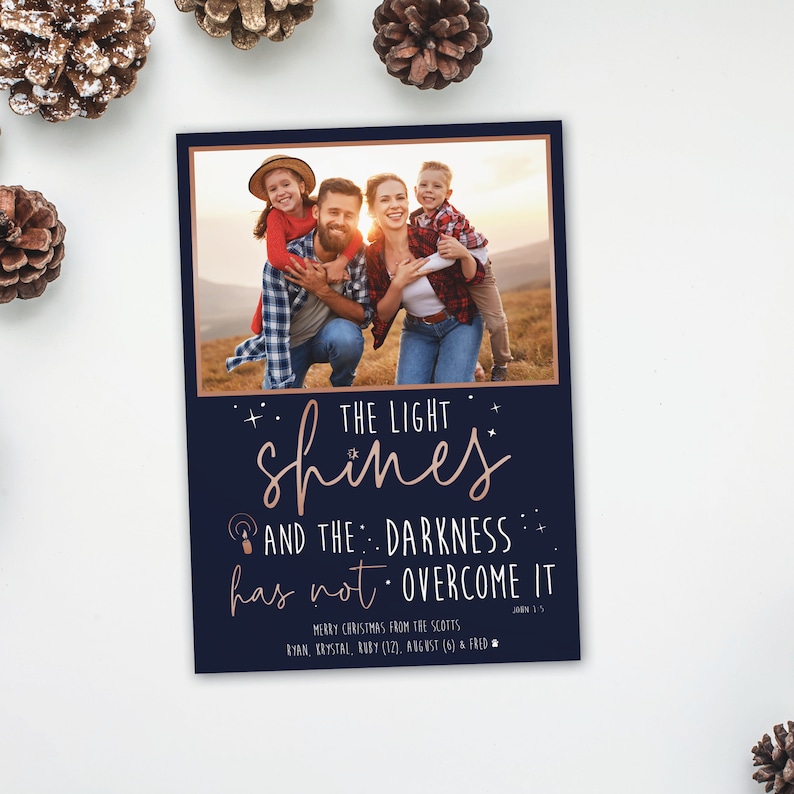 May include: A navy blue Christmas card with a family photo at the top. The card features the text "THE LIGHT shines AND THE DARKNESS has not OVERCOME IT" in a copper color. The card also includes the family's names and the Bible verse John 1:5.