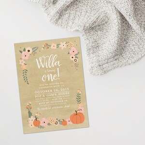 May include: A birthday invitation on kraft paper with floral and pumpkin accents. The text reads "Willa is turning one!" with event details. A gray knitted blanket is in the background.