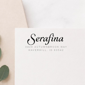 Puede incluir: Una tarjeta rectangular blanca con texto impreso en negro. El nombre Serafina está escrito en una fuente cursiva grande y elegante, seguido de la dirección 3428 Autumnbrook Way, Haverhill, ID 83542 en una fuente sans-serif simple.