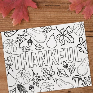 Puede incluir: Página para colorear en blanco y negro con la palabra "THANKFUL" rodeada de calabazas, hojas, bellotas, peras y otros elementos de la cosecha de otoño. Dos hojas de otoño rojas en el fondo.
