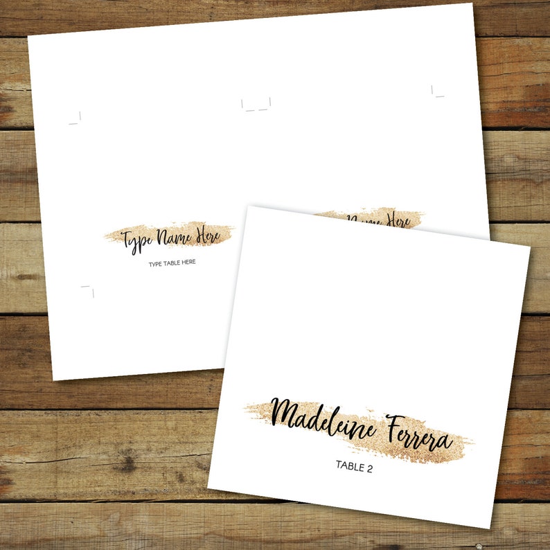 Puede incluir: Tarjetas de mesa blancas con detalles de pinceladas doradas con purpurina. Una tarjeta dice "Type Name Here" y "Type Table Here" en escritura negra. Otra tarjeta muestra "Madeleine Ferrera" y "Table 2".