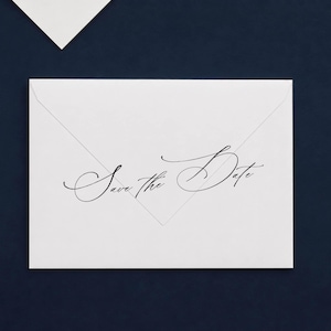May include: A white rectangular envelope featuring the text Save the Date printed in a black cursive script font across the center. The envelope flap is pointed and triangular, positioned against a dark blue background with a corner visible.