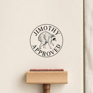 May include: A wooden rubber stamp featuring a circular design with a black ink illustration of a raccoon in the center. The text JIMOTHY is arched above the raccoon, and the word APPROVED is arched below it in a clean, black serif font.