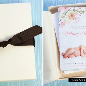 May include: A cream-colored gift box tied with a dark brown ribbon. Inside, a card announces "Whitney Elizabeth" with a photo of a newborn. Floral design in pink and green accents the card.