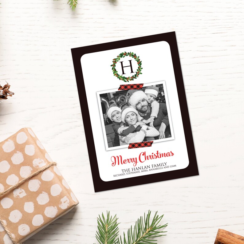 May include: A Christmas card with a black border, featuring a family photo and the words "Merry Christmas." The photo is secured with red and black plaid tape. A green wreath with the letter "H" is at the top.