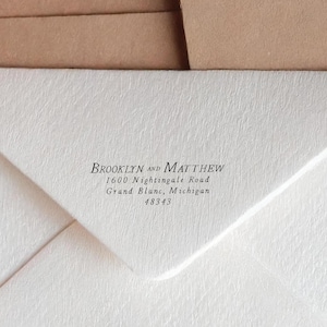 Op de afbeelding: Close-up van een witte envelop met de namen "Brooklyn and Matthew" in zwart script. Daaronder staat het adres: 1600 Nightingale Road, Grand Blanc, Michigan 48343. De envelop is gedeeltelijk open.