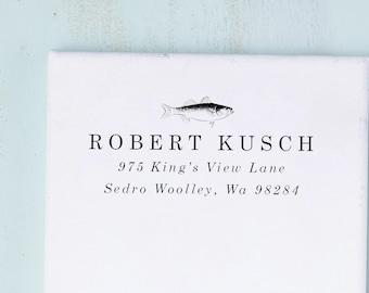 Può includere: Una tela rettangolare bianca con un'illustrazione a inchiostro nero di un pesce centrata sopra il testo ROBERT KUSCH, seguito dall'indirizzo 975 King's View Lane, Sedro Woolley, Wa 98284 stampato in un carattere serif nero.