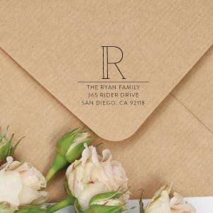 Pode incluir: Um envelope de papel castanho com um carimbo de tinta preta apresentando uma letra R grande, o texto THE RYAN FAMILY, 365 RIDER DRIVE e SAN DIEGO, CA 92118. O envelope é decorado com vários botões de rosa rosa pálido com caules e folhas verdes.
