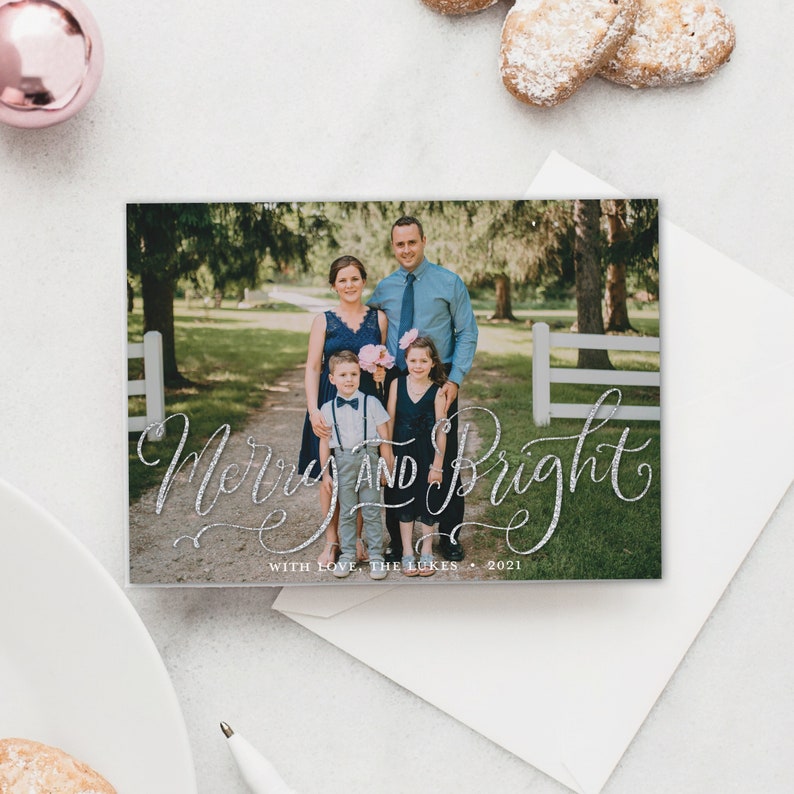 May include: A holiday card with a family portrait and the words "Merry and Bright" in silver glitter script. The family of four is standing on a path in front of a white fence. The card is on a white surface.