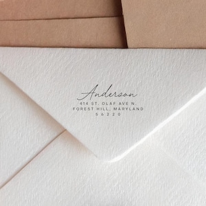 以下が含まれることがあります： Anderson, 414 St. Olaf Ave N., Forest Hill, Maryland, 56220と黒い文字が印刷された白い質感のある封筒のフラップ。封筒は、水平な折り目が見える茶色の紙の表面の前に配置されています。