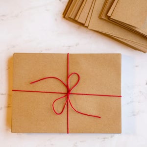 Puede incluir: Un paquete rectangular de tarjetas de papel marr&oacute;n atadas con hilo rojo, formando un lazo. Una pila de tarjetas similares se encuentra en la esquina superior derecha. Sobre una superficie de m&aacute;rmol blanco.