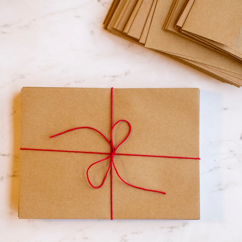 Puede incluir: Un paquete rectangular de tarjetas de papel marr&oacute;n atadas con hilo rojo, formando un lazo. Una pila de tarjetas similares se encuentra en la esquina superior derecha. Sobre una superficie de m&aacute;rmol blanco.