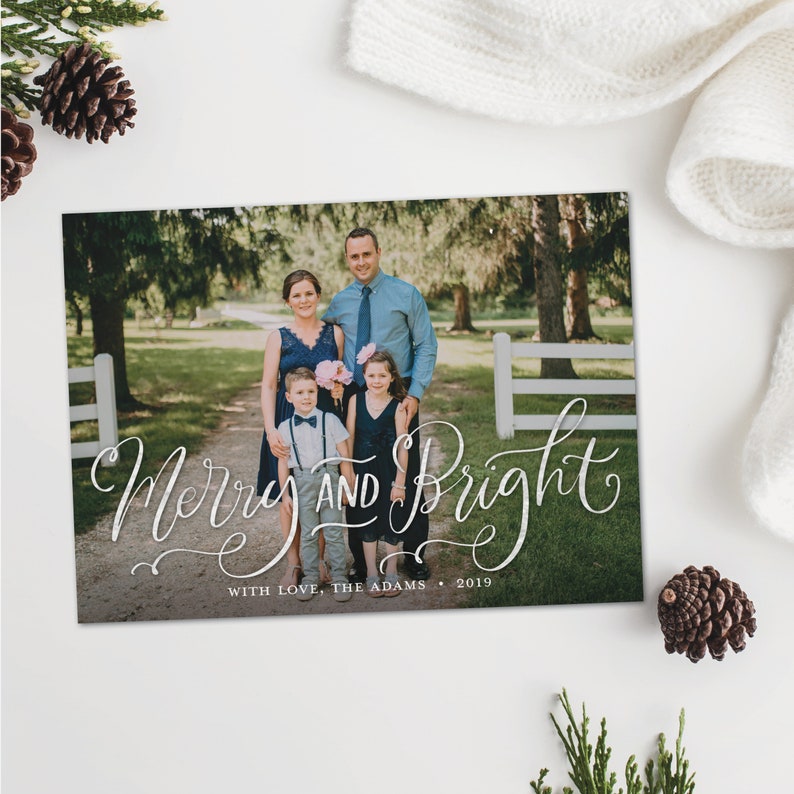 May include: A holiday card with a family portrait and the words "Merry and Bright" in elegant script. The family is standing on a path in front of a white fence and trees. The card includes the text "With Love, The Adams &bull; 2019".