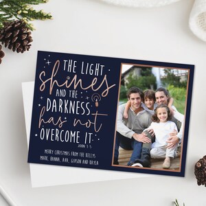 May include: A navy blue Christmas card with a family photo and the text "THE LIGHT shines AND THE DARKNESS has not OVERCOME IT." The card includes the names of the family members and the Bible verse John 1:5.