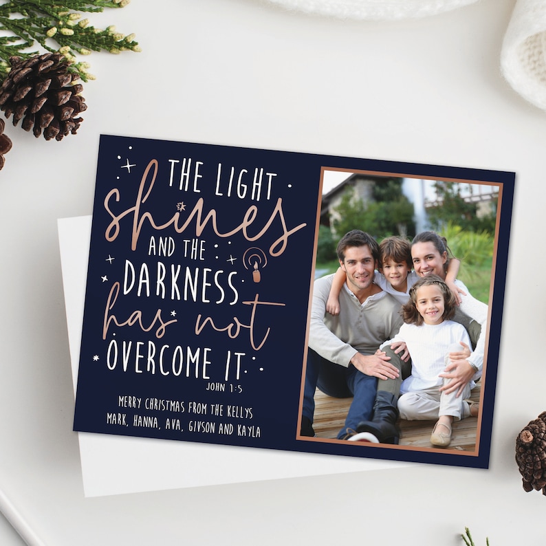 May include: A navy blue Christmas card with a family photo and the text "THE LIGHT shines AND THE DARKNESS has not OVERCOME IT." The card includes the names of the family members and the Bible verse John 1:5.