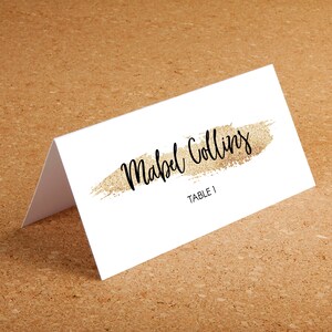Puede incluir: Una tarjeta de mesa blanca con el nombre "Mabel Collins" en escritura negra, subrayado por un trazo dorado brillante. El texto "TABLE 1" est&aacute; debajo. La tarjeta est&aacute; doblada y sobre una superficie marr&oacute;n.