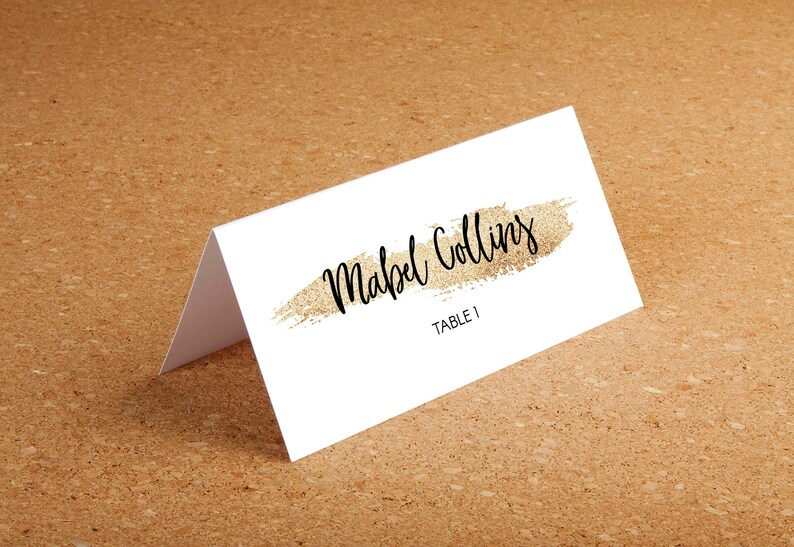 Puede incluir: Una tarjeta de mesa blanca con el nombre "Mabel Collins" en escritura negra, subrayado por un trazo dorado brillante. El texto "TABLE 1" est&aacute; debajo. La tarjeta est&aacute; doblada y sobre una superficie marr&oacute;n.