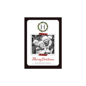 May include: A Christmas card with a black border and a white interior. The card features a family photo, a wreath with the letter "H", and the words "Merry Christmas". The family photo is secured with red and black plaid tape.