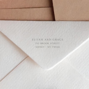 Pode incluir: Um envelope branco com superfície texturizada apresentando texto impresso em preto que diz ELIJAH AND GRACE, 193 BROOK STREET, SIDNEY, NY 79930. O envelope está posicionado contra um fundo castanho liso, exibindo um design limpo.
