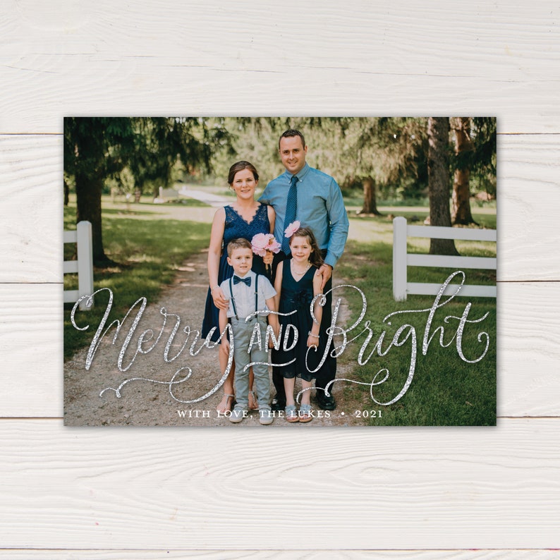 May include: A family portrait on a holiday card with the words "Merry and Bright" in silver script. The family of four is standing on a path, with trees and a white fence in the background. The card reads "With Love, The Lukes 2021".