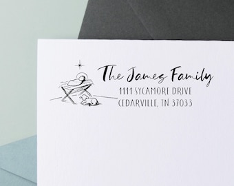 Op de afbeelding: Wit briefpapier met een zwarte inkt illustratie van een kerststal en de tekst "The James Family" en adresgegevens. Het briefpapier ligt op een lichtblauwe en donkergrijze achtergrond.