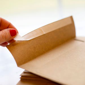 Puede incluir: Primer plano de una pila de sobres de papel marr&oacute;n. Una mano con esmalte de u&ntilde;as rojo despega la tira adhesiva de la solapa del sobre. Los sobres son de papel marr&oacute;n claro.