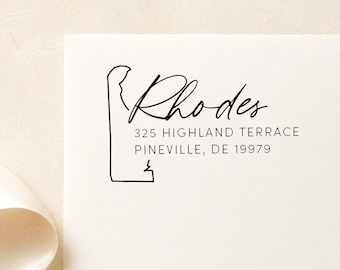 May include: A black ink stamp impression on white paper featuring a minimalist outline of the state of Delaware. The text reads Rhodes, 325 Highland Terrace, Pineville, DE 19979 in a combination of elegant script and clean sans-serif typography.