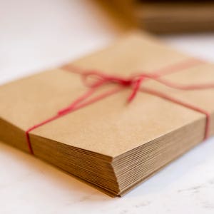 Puede incluir: Una pila de sobres de papel kraft marr&oacute;n, atados con hilo rojo. Los sobres son rectangulares y est&aacute;n apilados. El hilo est&aacute; atado con un lazo.