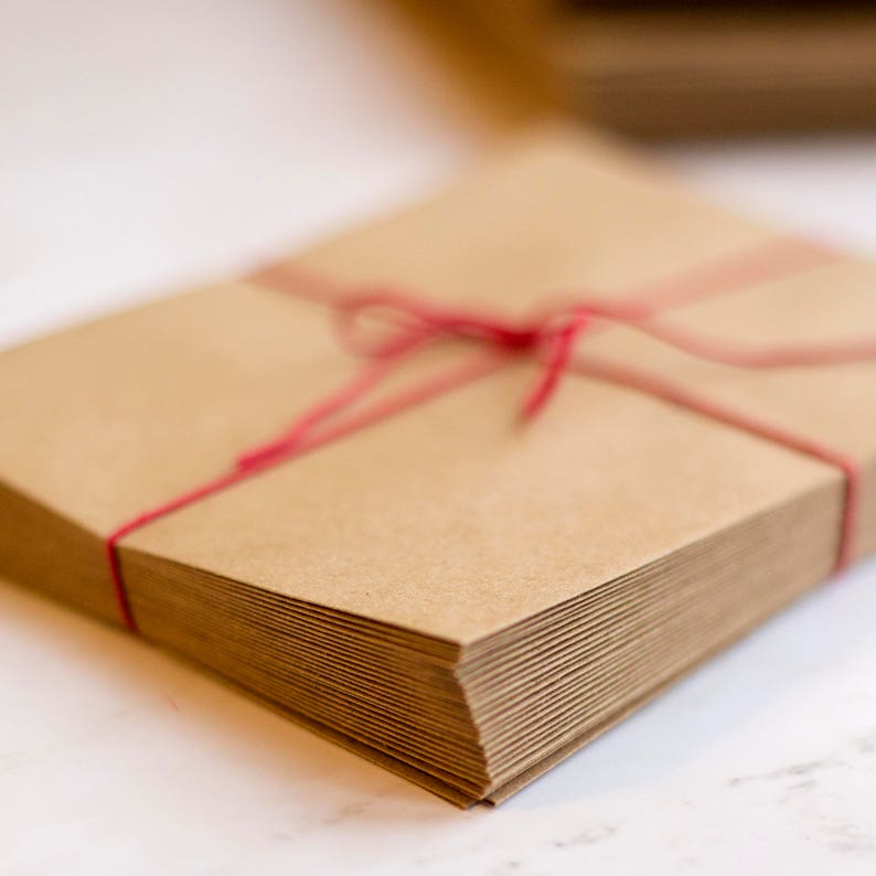 Puede incluir: Una pila de sobres de papel kraft marr&oacute;n, atados con hilo rojo. Los sobres son rectangulares y est&aacute;n apilados. El hilo est&aacute; atado con un lazo.