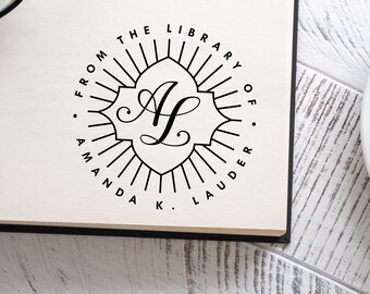 May include: A black and white custom library stamp with the text "FROM THE LIBRARY OF AMANDA K. LAUDER" encircling a decorative emblem with the letter "A" in the center. The stamp is on a white surface.