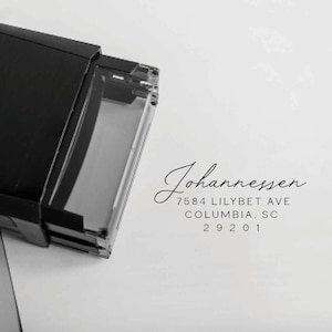 Op de afbeelding: Een zwarte adresstempel met de naam "Johannessen" in elegant schrift. Het adres is 7584 Lilybet Ave, Columbia, SC 29201. De stempel is gedeeltelijk zichtbaar, wat suggereert dat hij klaar is voor gebruik op papier.