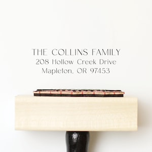 May include: A wooden address stamp with the text "THE COLLINS FAMILY" and address details. The stamp has a black handle and a rectangular imprint surface with red and black details. The background is plain white.