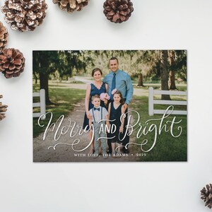 May include: A holiday card with a family portrait and the words "Merry and Bright" in elegant script. The family of four is standing on a path, surrounded by trees and a white fence. The card includes the text "With Love, The Adams &bull; 2019".
