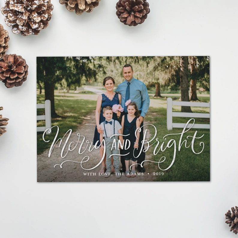 May include: A holiday card with a family portrait and the words "Merry and Bright" in elegant script. The family of four is standing on a path, surrounded by trees and a white fence. The card includes the text "With Love, The Adams &bull; 2019".