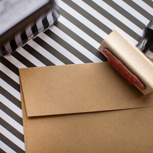 Puede incluir: Un sobre de papel kraft marr&oacute;n con un sello de madera y un sello a rayas blancas y negras. El sello tiene una almohadilla de tinta roja. El fondo es un patr&oacute;n de rayas blancas y negras.