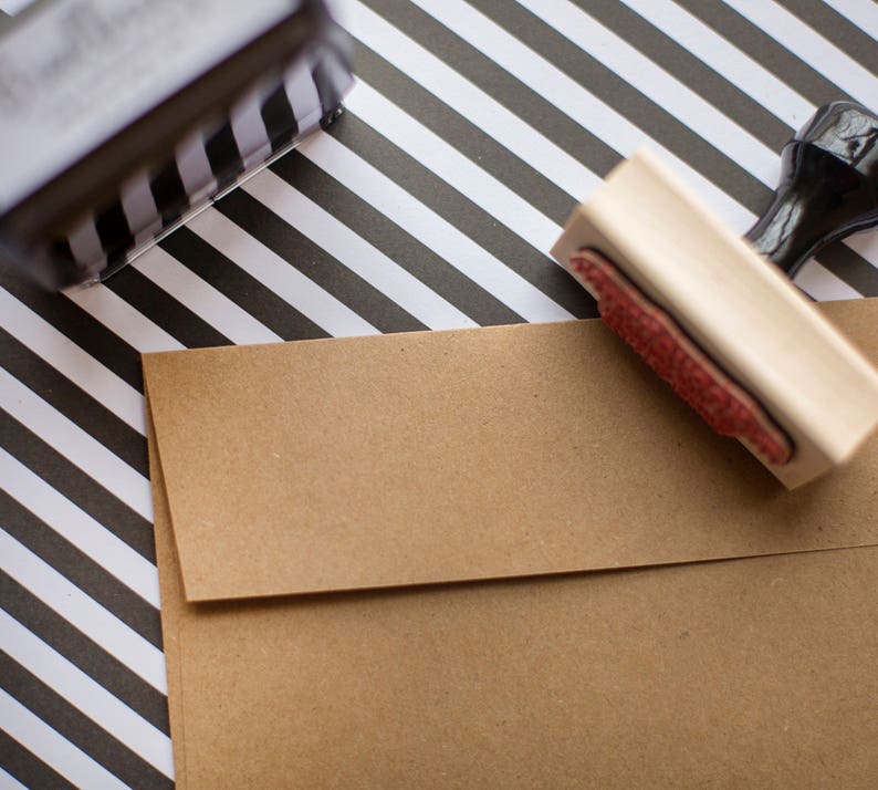 Puede incluir: Un sobre de papel kraft marr&oacute;n con un sello de madera y un sello a rayas blancas y negras. El sello tiene una almohadilla de tinta roja. El fondo es un patr&oacute;n de rayas blancas y negras.