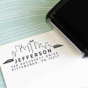 May include: A black ink stamp with a Pittsburgh skyline design, including the word "JEFFERSON" and an address. The stamp is partially pressed onto a white card, with a black stamp handle visible in the corner. The background is a light blue and white patterned surface.