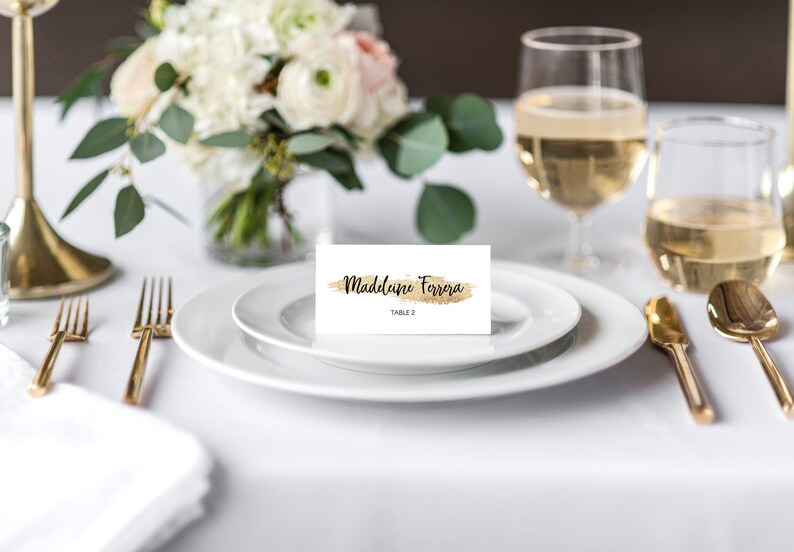 Puede incluir: Una mesa con un plato blanco, cubiertos dorados y una tarjeta con el nombre "Madeline Ferrera" y detalles dorados. Una copa de vino y un arreglo floral se encuentran en el fondo. La mesa est&aacute; preparada para un evento especial.