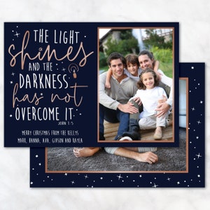 May include: A navy blue Christmas card with a family photo and text that reads "THE LIGHT shines AND THE DARKNESS has not OVERCOME IT." The card has a copper-colored border and star accents. The family photo shows a smiling family of four.