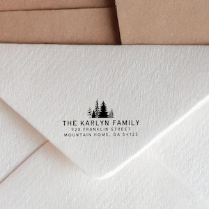 Könnte beinhalten: Weißer Umschlag mit schwarzer Waldsilhouette und Text. Der Text lautet "THE KARLYN FAMILY" über der Adresse "928 FRANKLIN STREET MOUNTAIN HOME, GA 54123". Der Umschlag besteht aus strukturiertem Papier.