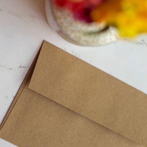 Puede incluir: Dos sobres de papel marr&oacute;n, uno ligeramente superpuesto al otro, est&aacute;n colocados en diagonal. Los sobres tienen una superficie texturizada y est&aacute;n sobre un fondo de m&aacute;rmol blanco. La esquina superior de los sobres es visible.