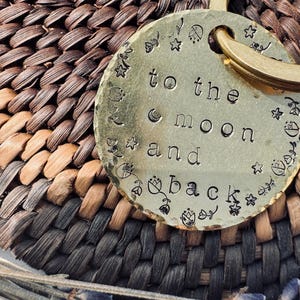Può includere: Un portachiavi in metallo dorato con un'etichetta rotonda che recita "to the moon and back". L'etichetta è decorata con piccole stelle e motivi floreali.