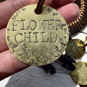 Può includere: Un portachiavi in ​​ottone con un'etichetta rotonda che dice "FLOWER CHILD" in maiuscolo. L'etichetta è decorata con un motivo floreale e funghi.