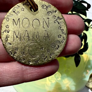 Può includere: Un portachiavi in metallo dorato con le parole "MOON MAMA" incise. Il portachiavi è decorato con una luna crescente e stelle.