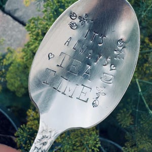 May include: A silver spoon with the phrase "IT'S ALWAYS TEA TIME" stamped on the bowl. The spoon is decorated with floral and heart motifs. The handle has a floral design. The background is blurred green foliage.