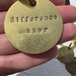 Può includere: Un portachiavi in metallo dorato con un'etichetta rotonda su cui è scritto "difference maker".