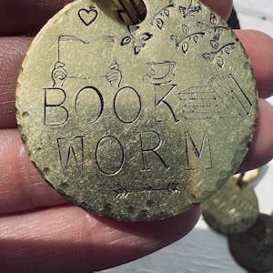 Può includere: Un portachiavi in ​​ottone con finitura martellata che presenta il testo "BOOK WORM" con un piccolo cuore, un libro, una tazza di tè e una pila di libri.