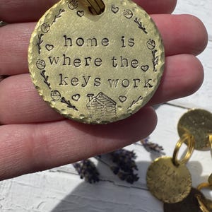 Può includere: Un portachiavi in ​​ottone con un'etichetta rotonda su cui è scritto "home is where the keys work". L'etichetta è decorata con una piccola casa, cuori e motivi floreali.