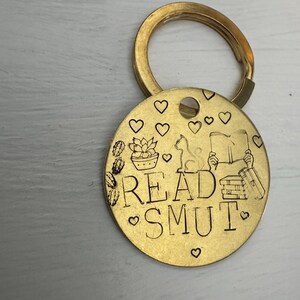 Può includere: Un portachiavi in metallo dorato con un'etichetta rotonda che dice "READ SMUT" in un font stilizzato. L'etichetta è decorata con cuori, un gatto, una pianta in vaso e una pila di libri.