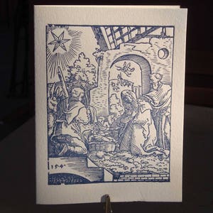 May include: A blue and white Christmas card featuring a detailed illustration of the Nativity scene. The artwork depicts Mary, Joseph, and baby Jesus, with a star shining above. The card is held by a metal clip.